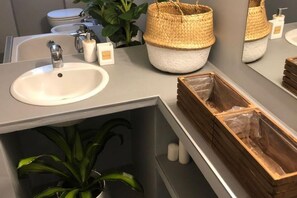 Une salle de bain avec un lavabo blanc, un miroir, un grand panier en osier et une boîte en bois.