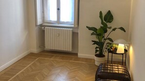 Une pièce avec un radiateur, une fenêtre avec des rideaux, une plante en pot et une lampe sur une table d’appoint.