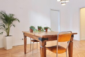 Una mesa de comedor de madera con sillas, plantas en macetas y una lámpara colgante en una habitación con suelo de madera y paredes blancas.