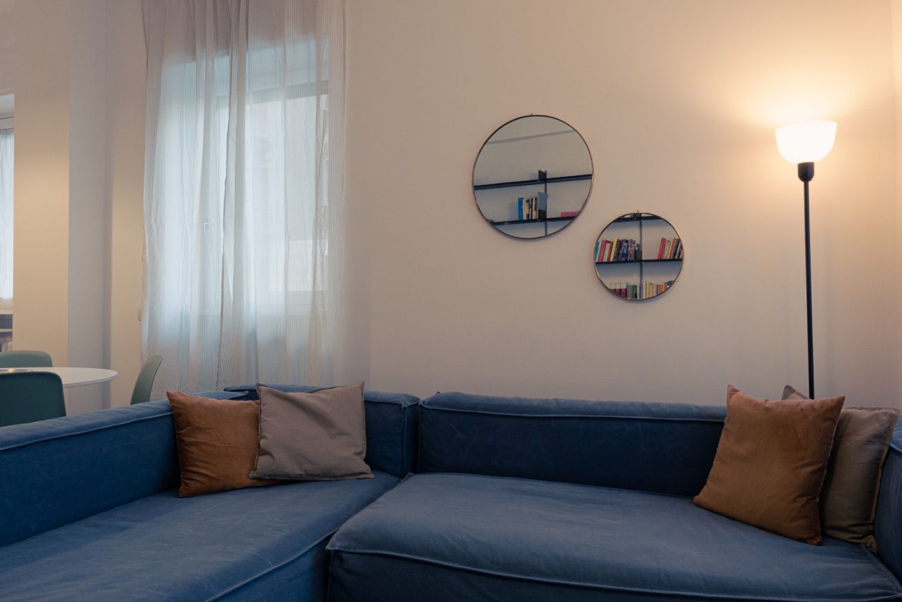 Un canapé bleu avec deux coussins, un miroir rond et une lampe sur pied dans une pièce dotée d’une fenêtre et d’une bibliothèque.