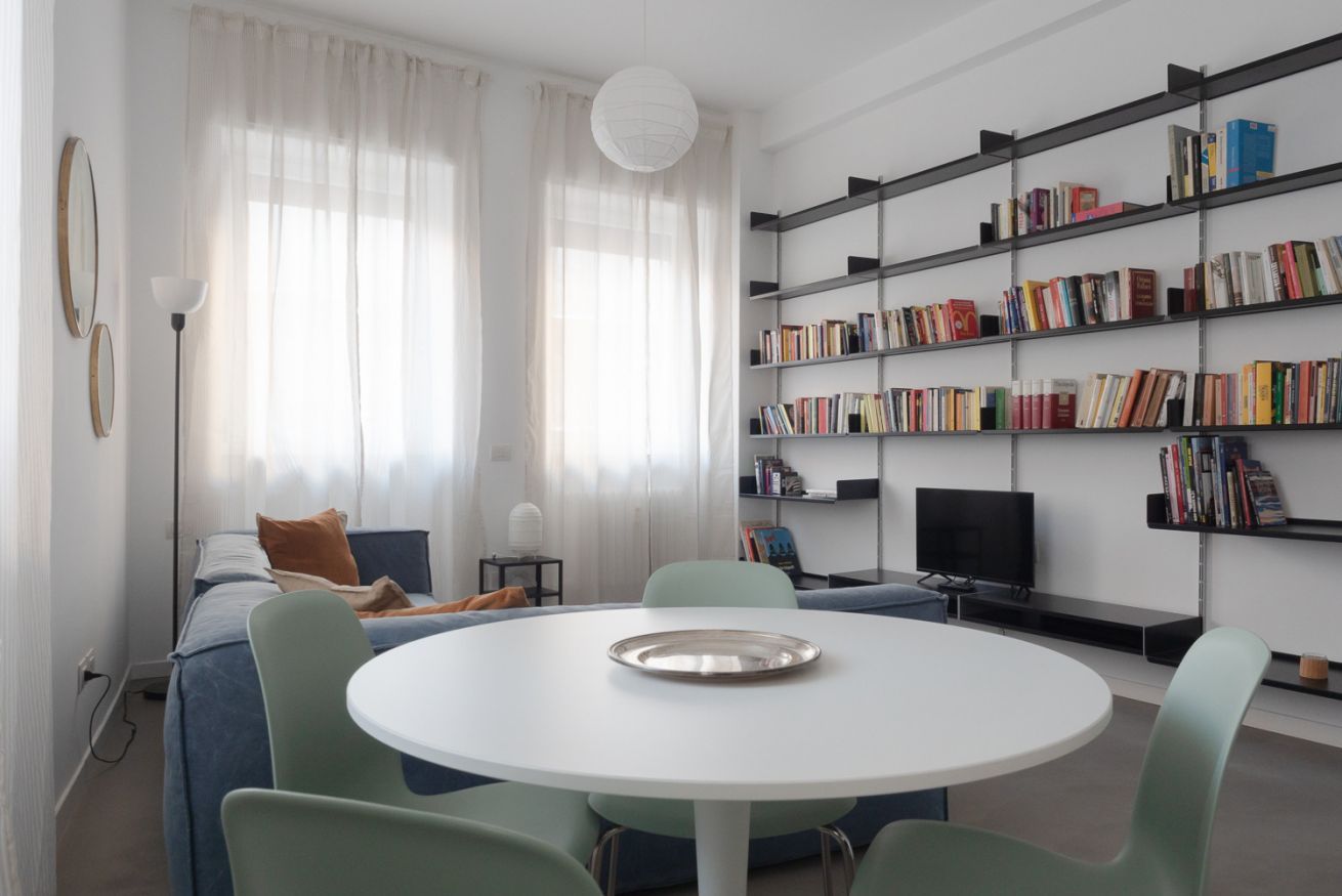 A modern living room with a round table, green chairs, a sofa, a bookshelf, and a television.