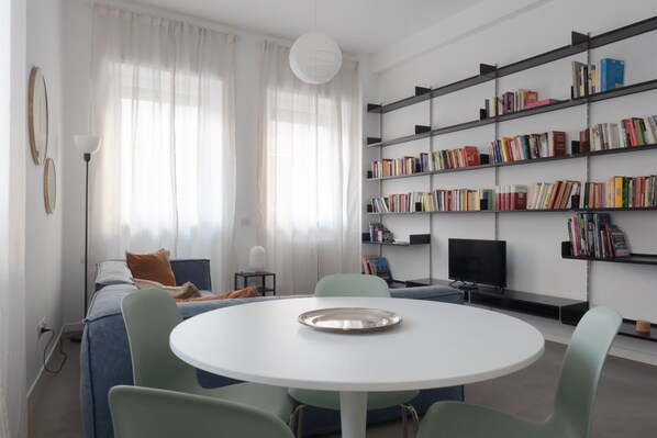 A modern living room with a round table, green chairs, a sofa, a bookshelf, and a television.
