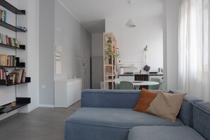 A modern living room with a blue sofa, a bookshelf, a dining area with a table and chairs, and a kitchen in the background.
