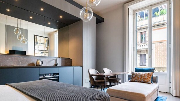 A modern hotel room with a bed, a desk, a chair, a sofa, a large window, and a kitchen area. - Paganini boutique apartment (Milano)