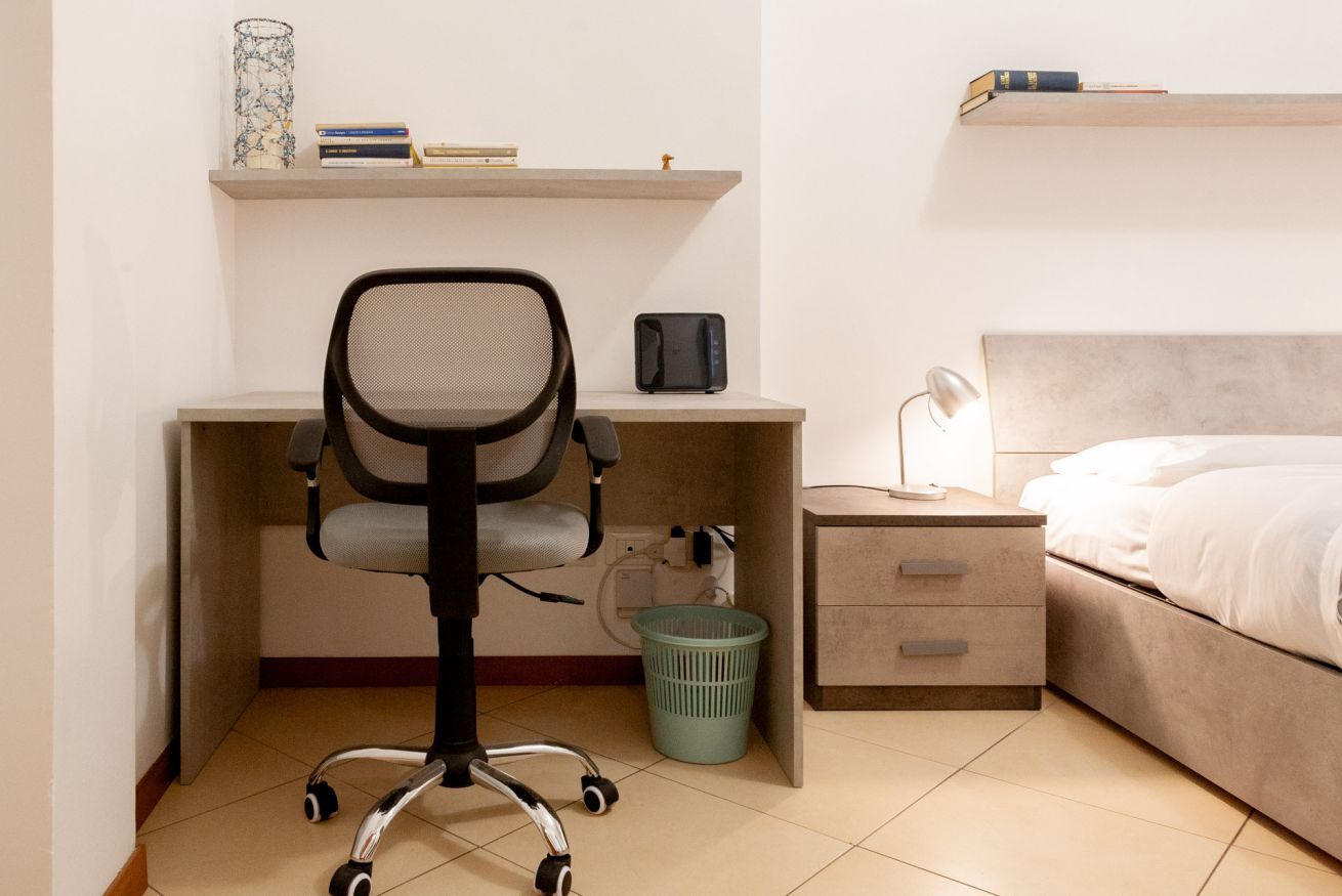 Una camera da letto con un letto, una scrivania con sedia, un comodino, una lampada e una mensola a parete con libri e un oggetto decorativo.