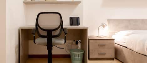 Un dormitorio con cama, escritorio y silla, mesita de noche, lámpara y estante en la pared con libros y un objeto decorativo.