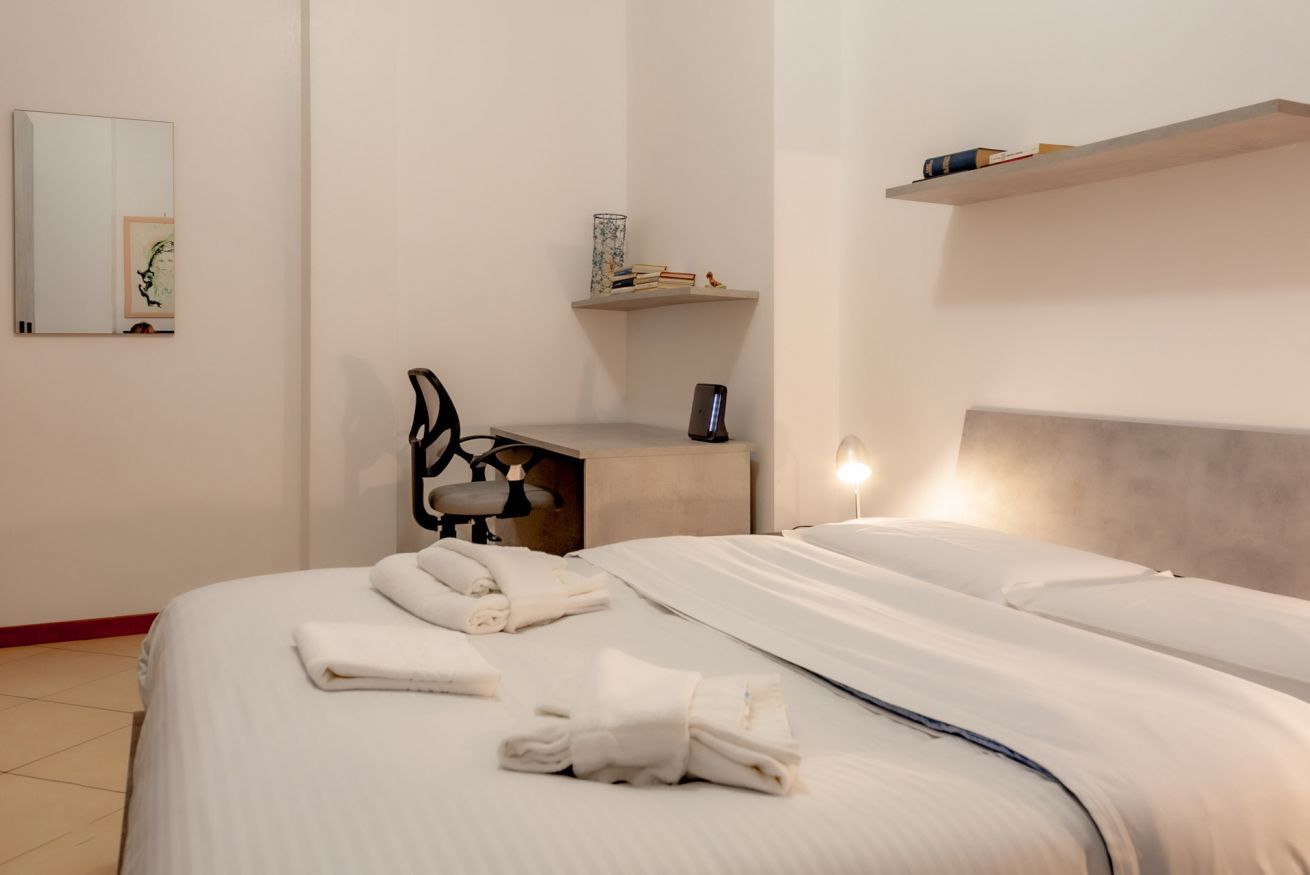 A hotel room with a bed, a desk, a chair, a mirror, and a shelf with books and a vase.