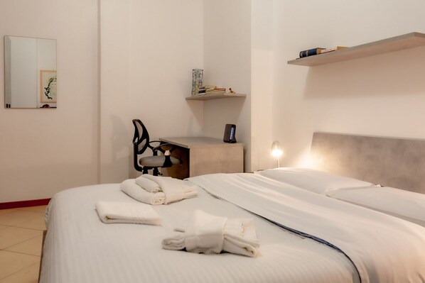 Une chambre d’hôtel avec un lit, un bureau, une chaise, un miroir et une étagère contenant des livres et un vase.