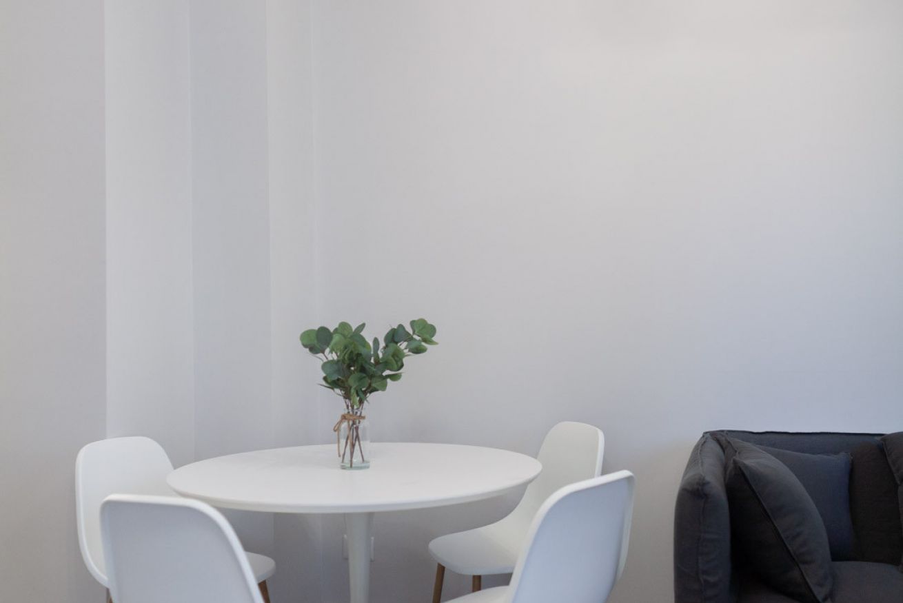 Een ronde witte tafel met vier witte stoelen, een kleine plant in een glazen vaas en een donkergrijze bank.