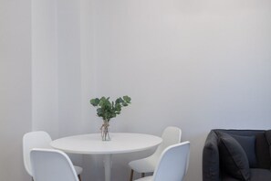 Une table ronde blanche, accompagnée de quatre chaises blanches, une petite plante dans un vase en verre et un canapé gris foncé.