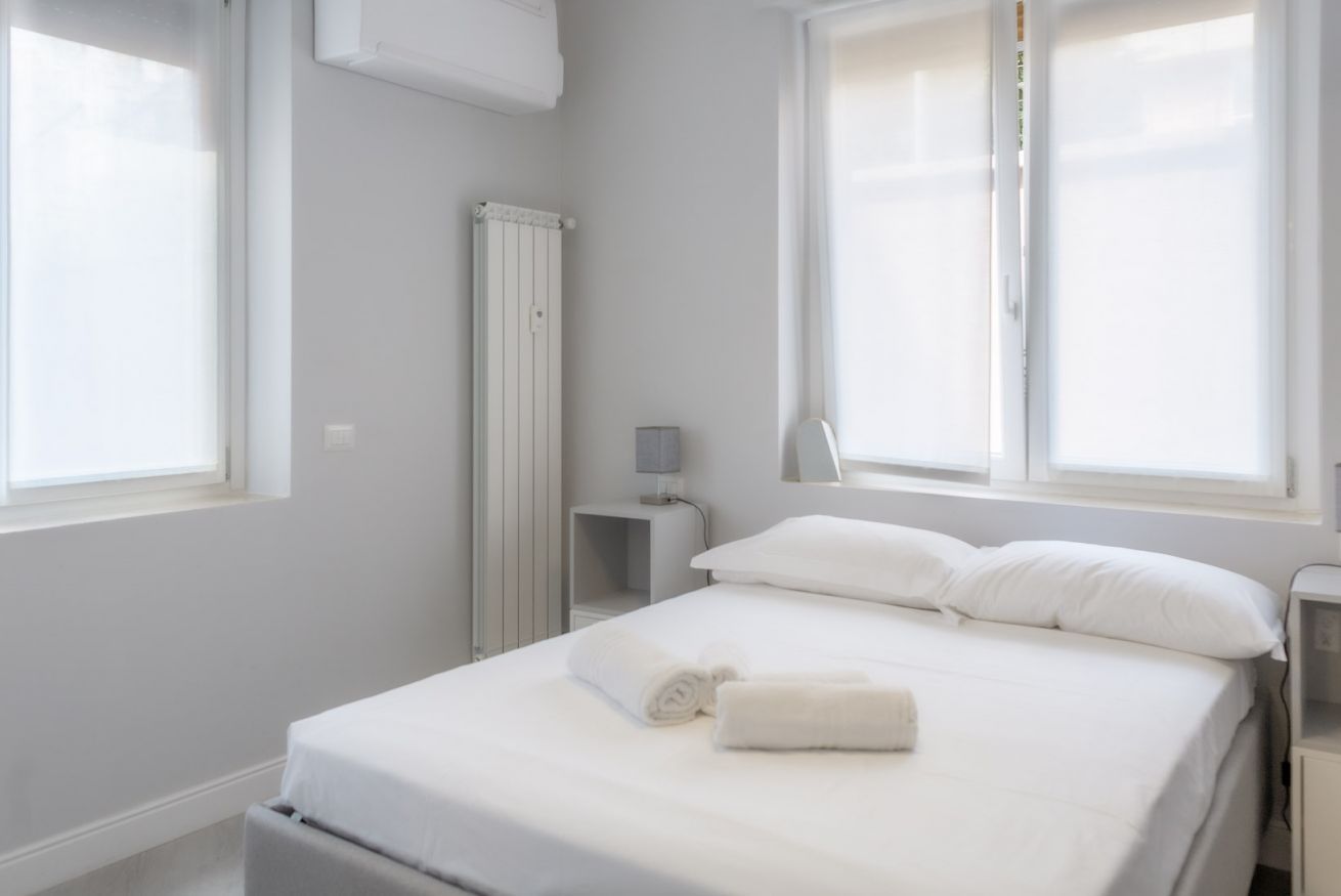 Habitación de hotel minimalista con cama, un radiador, una mesita de noche y una ventana.