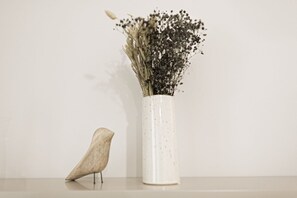 A white vase with a speckled pattern containing dried flowers and a small bird figurine on a reflective surface against a plain wall.