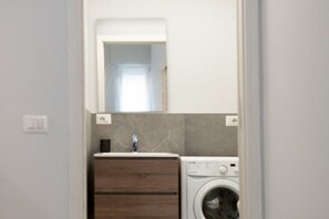 Une petite buanderie équipée d’un lave-linge, d’un évier et d’un miroir au-dessus du plan de travail.