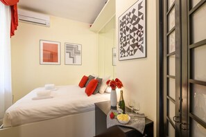 Une chambre d’hôtel avec un lit, une table de chevet sur laquelle se trouvent une bouteille, des verres et un bol de fruits. Deux œuvres d’art encadrées ornent le mur.