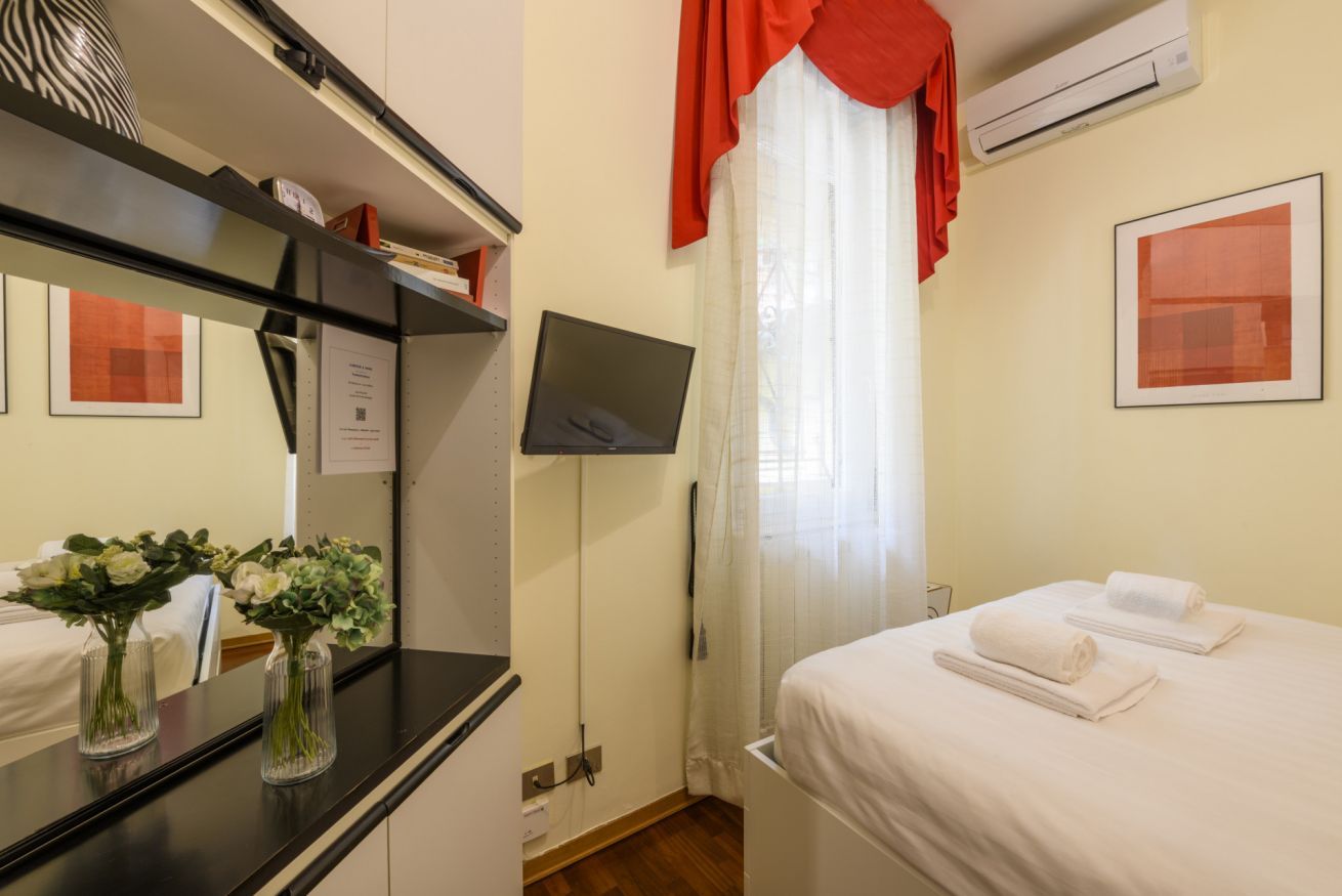 A hotel room with a bed, a desk with a mirror, a flat-screen TV, and a window with curtains.