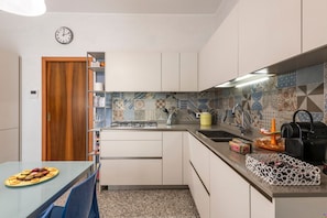 A compact kitchen with a wooden door, a clock, a tiled backsplash, and a dining area with a table set for two. - Stromboli, 22 – Elegante appartamento (Milano)
