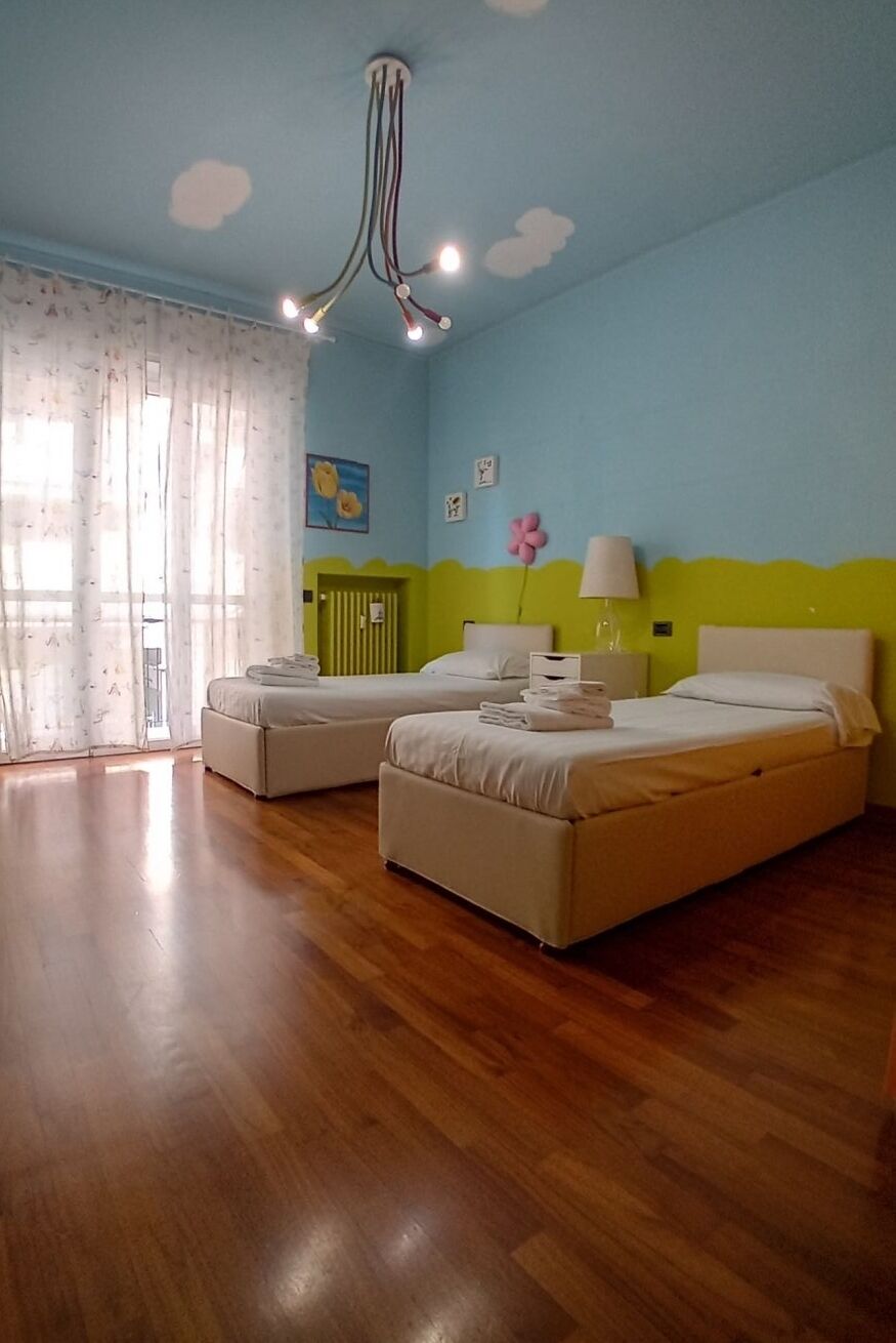 Una stanza con due letti, pavimento in legno, soffitto decorato con nuvole e una finestra con tende.
