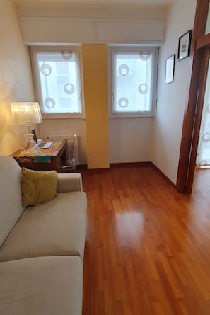 A small, furnished room with a wooden floor, a sofa, a table with a lamp, and two windows with curtains. - Stromboli, 22 – Elegante appartamento (Milano)