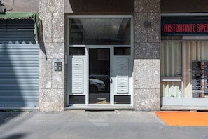 A building with a closed shutter on the left, a glass door entrance with a car inside, and a restaurant sign on the right. - Stromboli, 22 – Elegante appartamento (Milano)