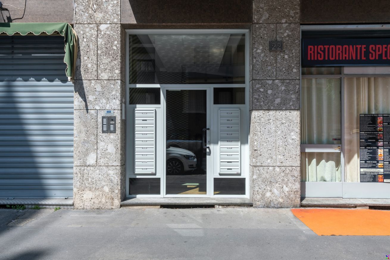 Un edificio con una persiana chiusa a sinistra, un ingresso con porta in vetro e un'auto all'interno, e un cartello di un ristorante a destra.