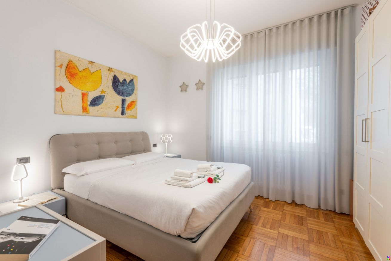 Quarto com cama, mesa de cabeceira, luminária, quadro e janela com cortinas.