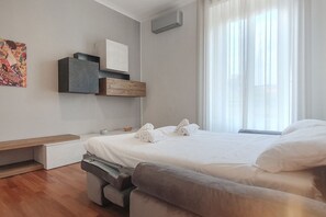 Quarto de hotel moderno com cama, mesas de cabeceira, um banco e uma janela grande com cortinas leves.