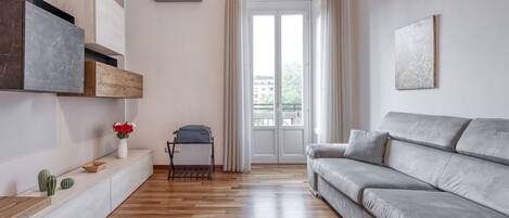 A modern living room with a grey sofa, wooden flooring, a white console table with decorative items, and a sliding glass door leading to an outdoor area.