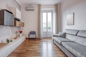 Uma sala de estar moderna com um sofá cinzento, piso de madeira, um console branco com objetos decorativos e uma porta de vidro deslizante que dá para uma área externa.