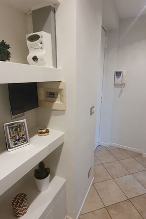 Um corredor com paredes brancas, chão em mosaico, televisão montada na parede, termostato, telefone e uma foto emoldurada em uma prateleira.