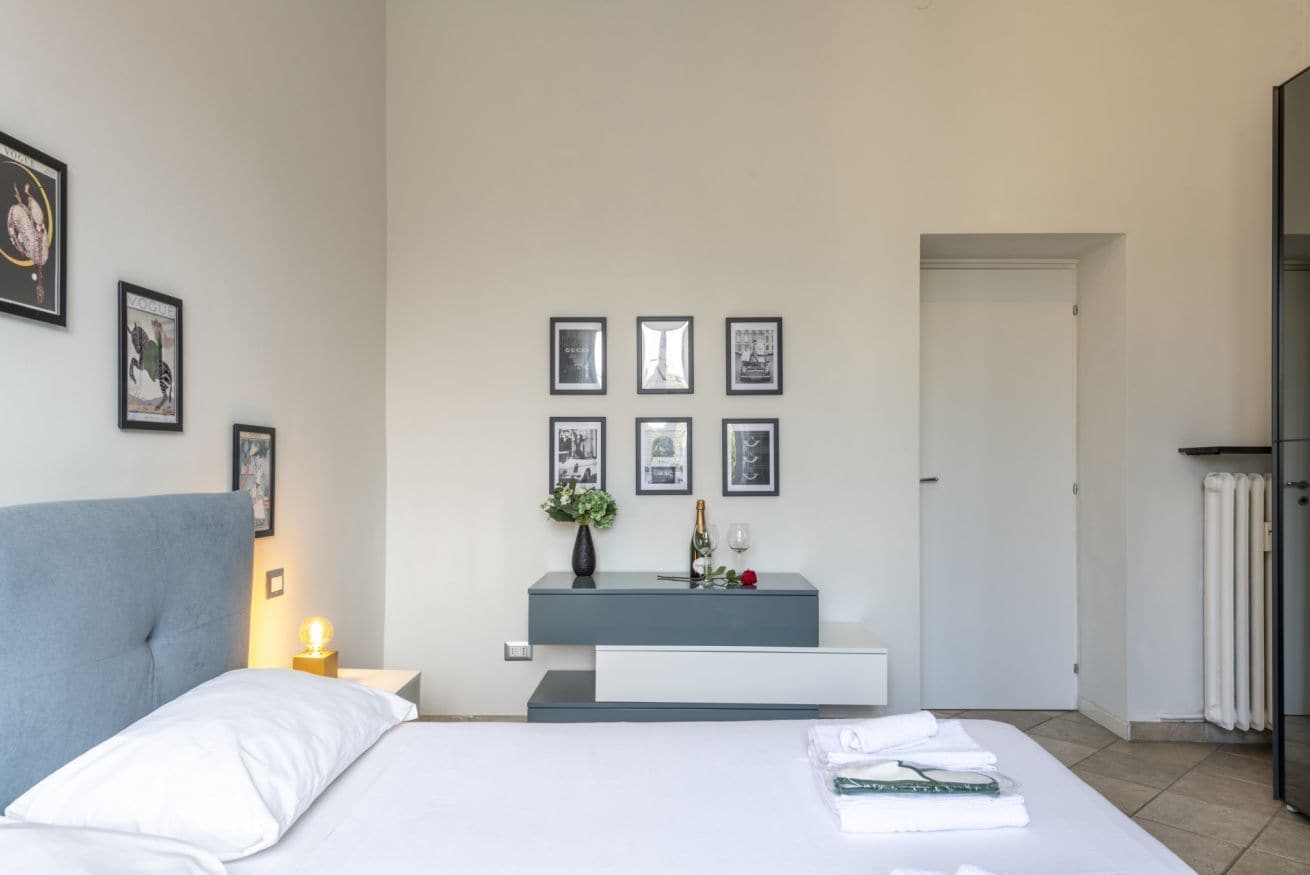 A bedroom with a bed, bedside table, wall-mounted shelf, framed pictures, and a door.
