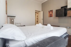 A hotel room with a bed, a chair, a radiator, a painting, and a TV on the wall.