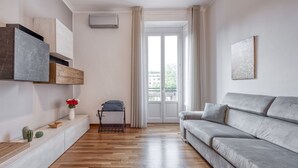 A modern living room with a grey sofa, wooden flooring, a white console table with decorative items, and a sliding glass door leading to an outdoor area. - Venini 14 - Tra Centrale e Buenos Aires (Milano)