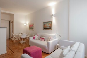 A modern living room with a white sofa, a pink ottoman, a dining table with chairs, and two wall-mounted lamps.