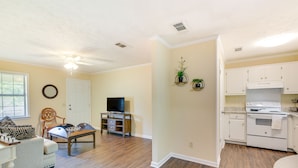 A living room with a sofa, a coffee table, and a TV stand. There is a kitchen with white cabinets and a stove. - Bring the Whole Alumni Crew Close to Ole Miss (Oxford)
