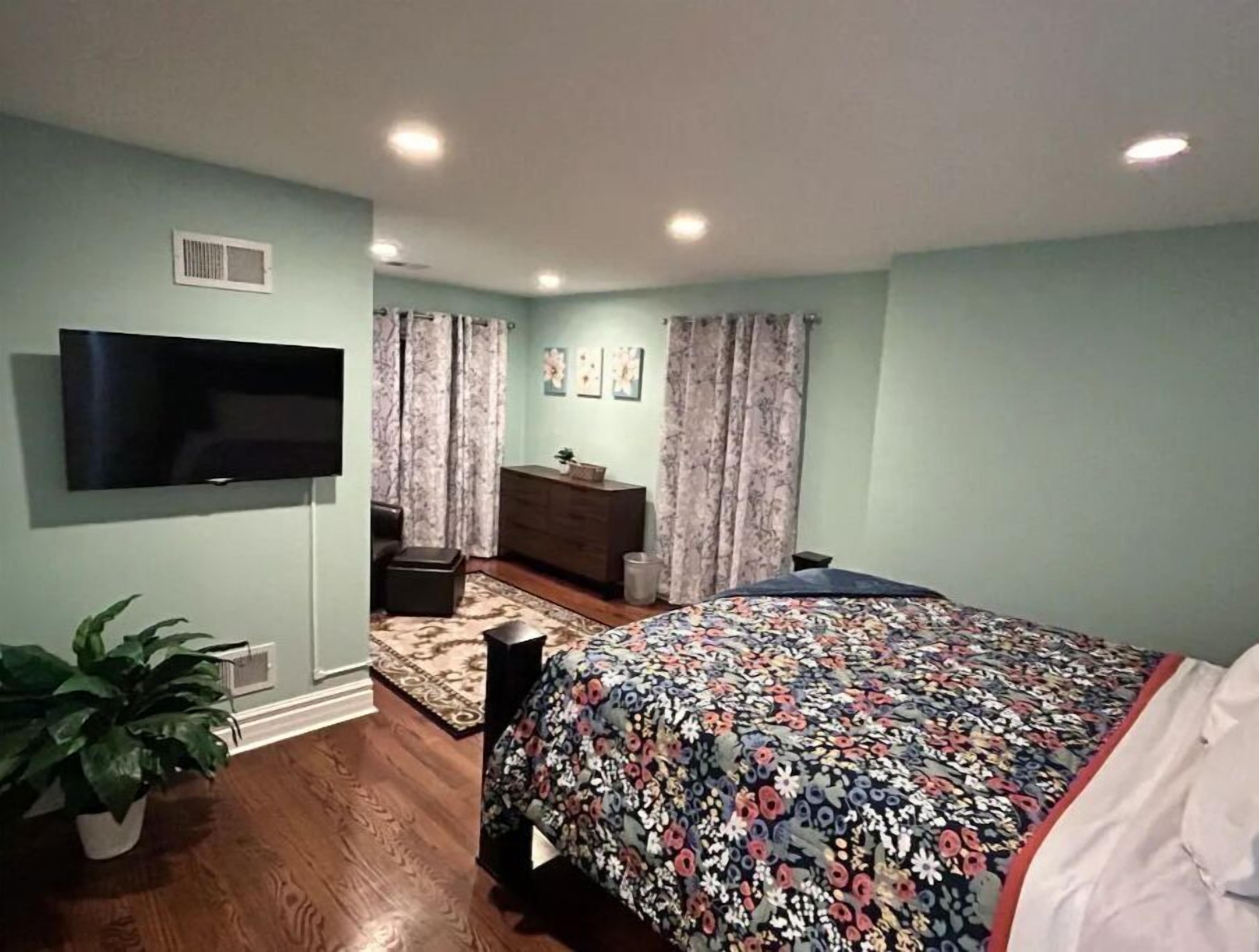A bedroom with a bed, a TV mounted on the wall, a dresser, a plant, and a window with curtains.