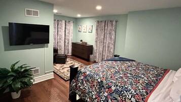 A bedroom with a bed, a TV mounted on the wall, a dresser, a plant, and a window with curtains.