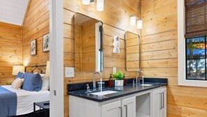 Interior de una cabaña de madera con un espejo grande, una cama con ropa de cama azul, un lavabo con encimera y una ventana con persianas.