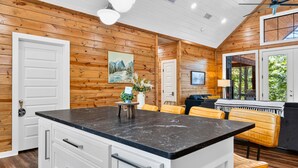 A kitchen with wooden walls, a central island with a black countertop, white cabinets, and a dining area with yellow chairs.