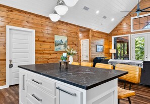 A kitchen with wooden walls, a central island with a black countertop, white cabinets, and a dining area with yellow chairs.