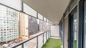 Ein schmaler Balkon mit einem grünen Kunstrasenteppich, großen Fenstern mit Blick auf umliegende Gebäude und einem Metallgeländer.