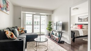 Ein modernes Wohnzimmer mit einer Couch, einem Couchtisch und einem Fernseher. Ein großes Fenster bietet Blick auf Gebäude.