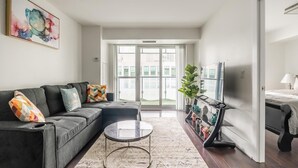 Ein modernes Wohnzimmer mit einem grauen Ecksofa, einem runden Couchtisch und einem großen Fenster mit Blick ins Freie.