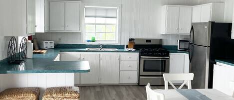 Une cuisine moderne avec des meubles blancs, un îlot central, un coin repas avec des chaises blanches et une grande fenêtre qui laisse entrer la lumière naturelle.