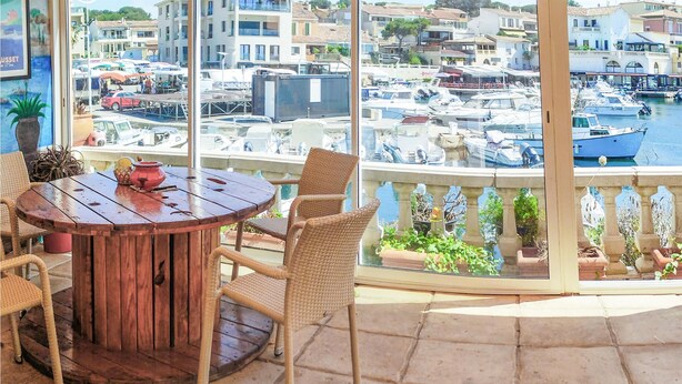 A round wooden table with wicker chairs, a potted plant, and a red pot on a balcony overlooking a marina with boats and buildings.