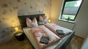 Ein ordentlich bezogenes Bett mit rosa Bettwäsche und zwei Kissen, eine Nachttischlampe und ein kleiner Mülleimer.