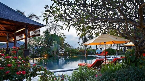 Un área de piscina tranquila con sillones de descanso y sombrillas, rodeada de exuberante vegetación y un jardín cuidado.