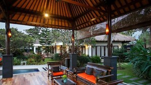 Un espacio de estar al aire libre cubierto, con mobiliario de madera y una piscina al fondo.