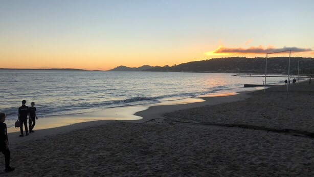 Una playa con un mar tranquilo, un atardecer de fondo y algunas personas caminando por la orilla.