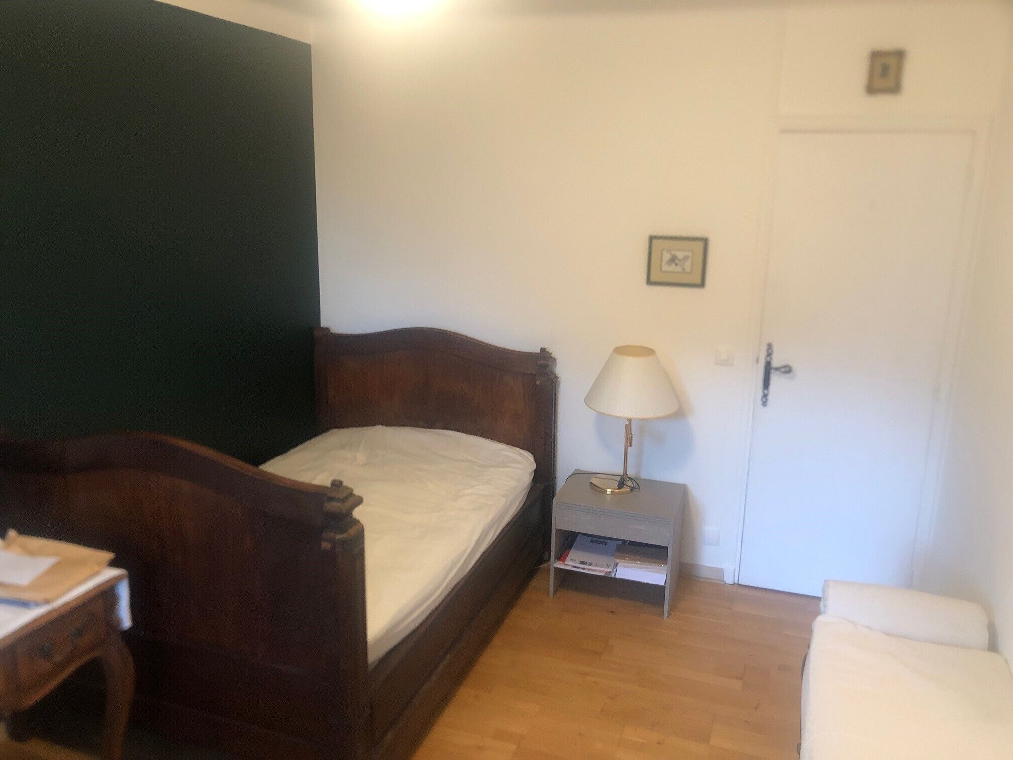A small, neatly arranged bedroom with a wooden bed, a bedside table with a lamp, a small shelf with books, and a door with a picture frame.