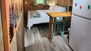 Interior rústico de una cabaña con una cama, una mesita, un refrigerador y un cuadro de árboles.